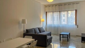 Una sala de estar moderna con un sofá, una mesa de comedor y sillas. Hay una lámpara y una mesita. La habitación tiene un ventanal con cortinas.