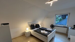 A bedroom with a bed, bedside table, lamp, fan, and a window with a view of a house. - Holiday Home 'Almir' with Private Terrace, Private Garden and Wi-Fi (Haselünne)