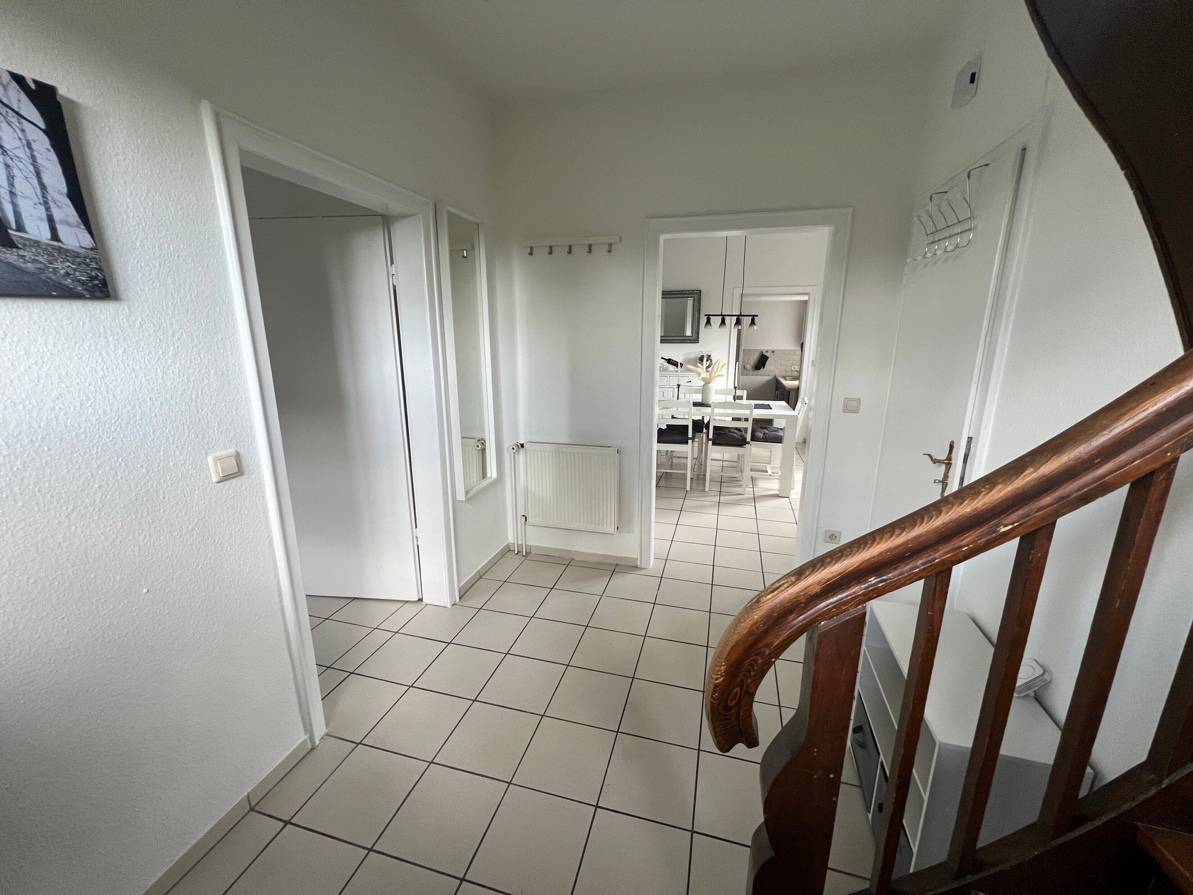 Een hal die uitkomt op een keuken met witte muren, een betegelde vloer en een houten trap.
