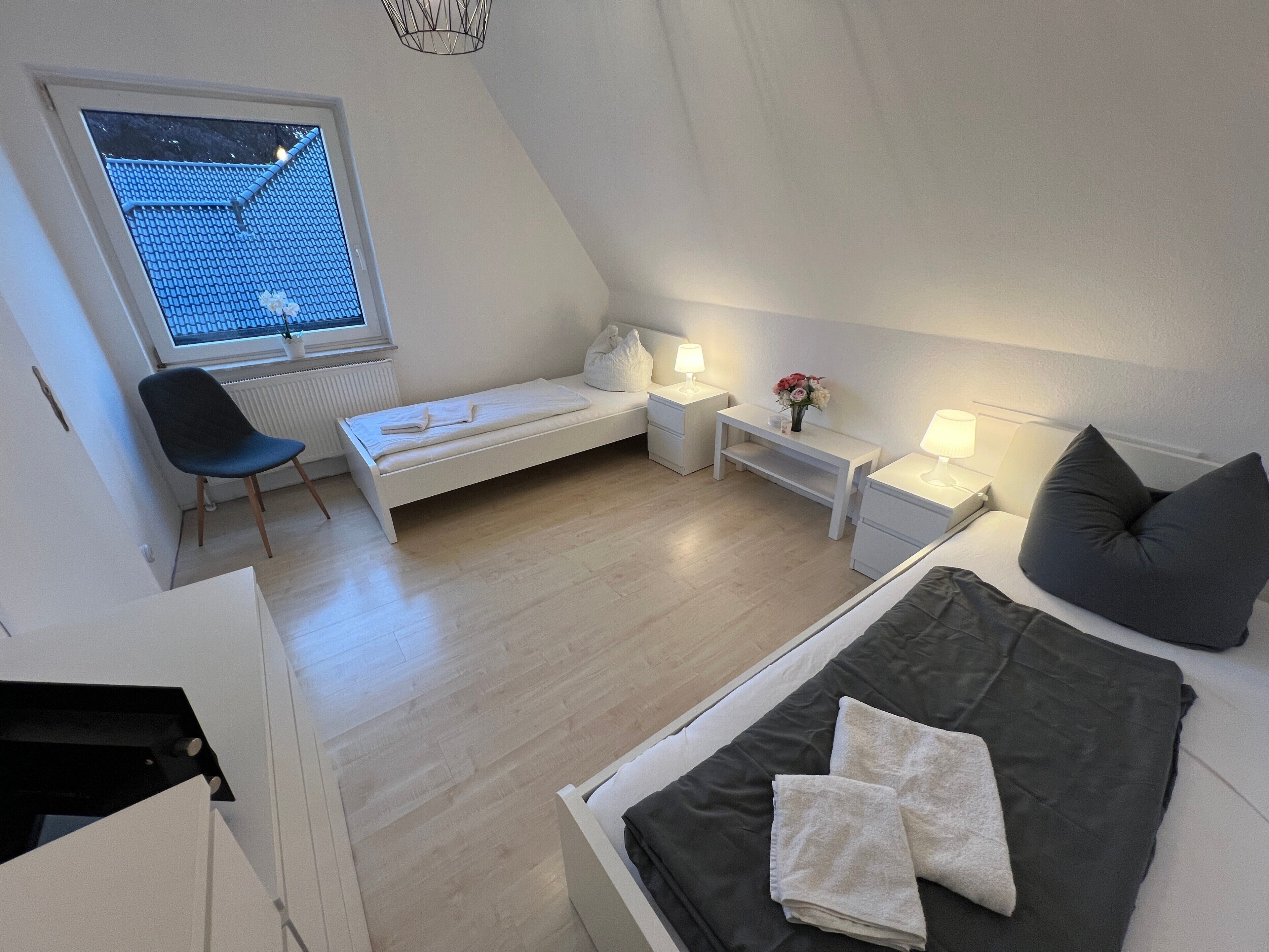 A modern, compact room with a bed, a chair, a small table, and a window with a view of the roof.