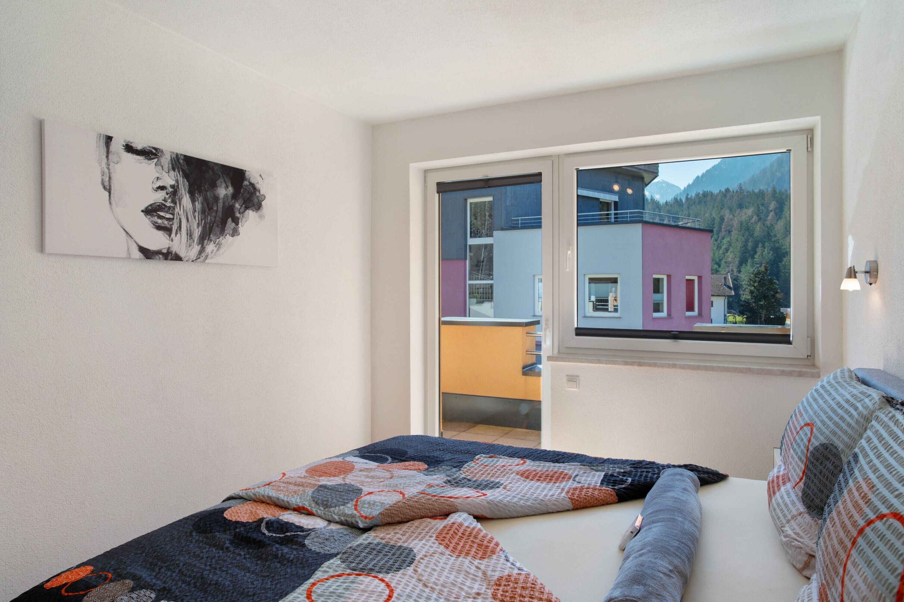 A bedroom with a bed, a window with a view of buildings, and a painting on the wall.