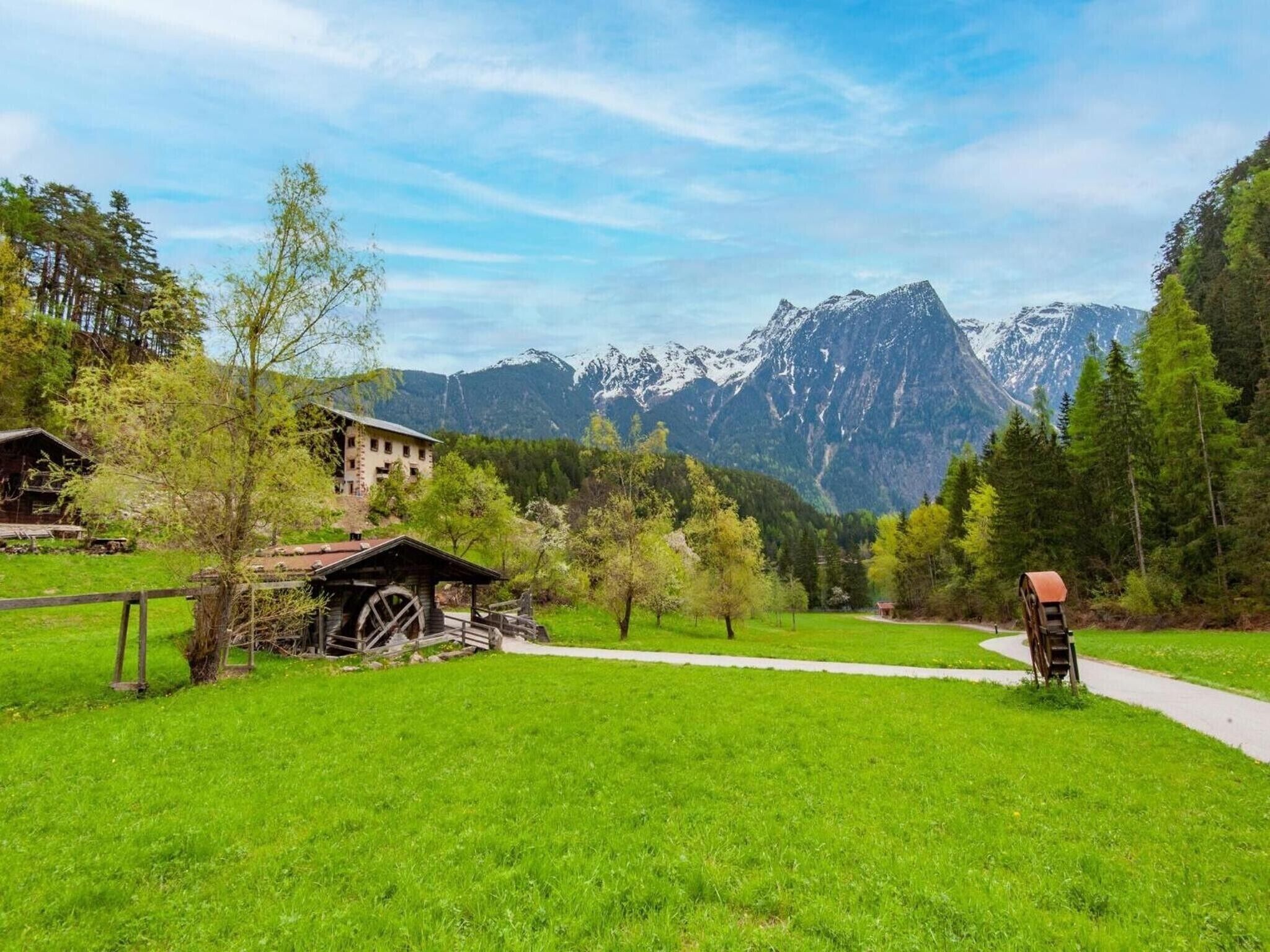 Paisagem montanhosa com uma cabana de madeira, um moinho de água e um caminho serpenteando por um campo verde.