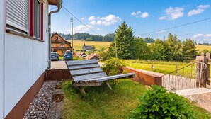 Uma propriedade residencial com deck de madeira, entrada com calçamento e vista para paisagem rural com casas e árvores.