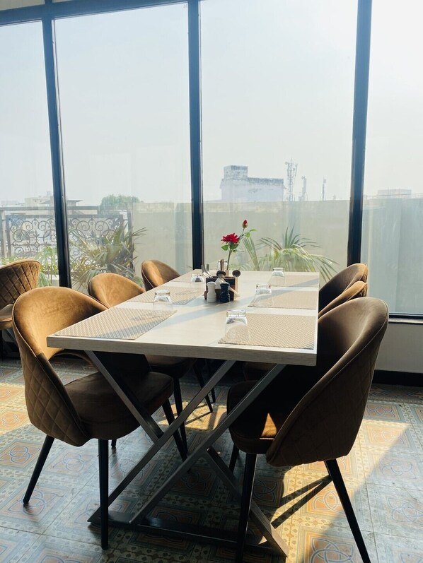 A dining area with a table set for six, surrounded by six chairs, with a view of buildings through large windows. - Hotel Glorious Grand (Varanasi)
