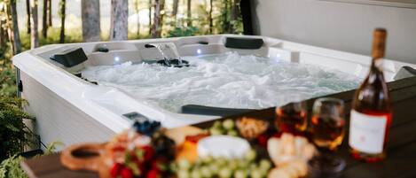 Un bain à remous extérieur, entouré de verdure, avec une table remplie de nourriture et de boissons devant.