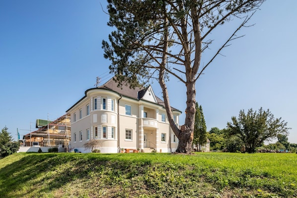 Un grand immeuble à plusieurs étages, avec un arbre remarquable qui se dresse devant, entouré d’une zone verdoyante.