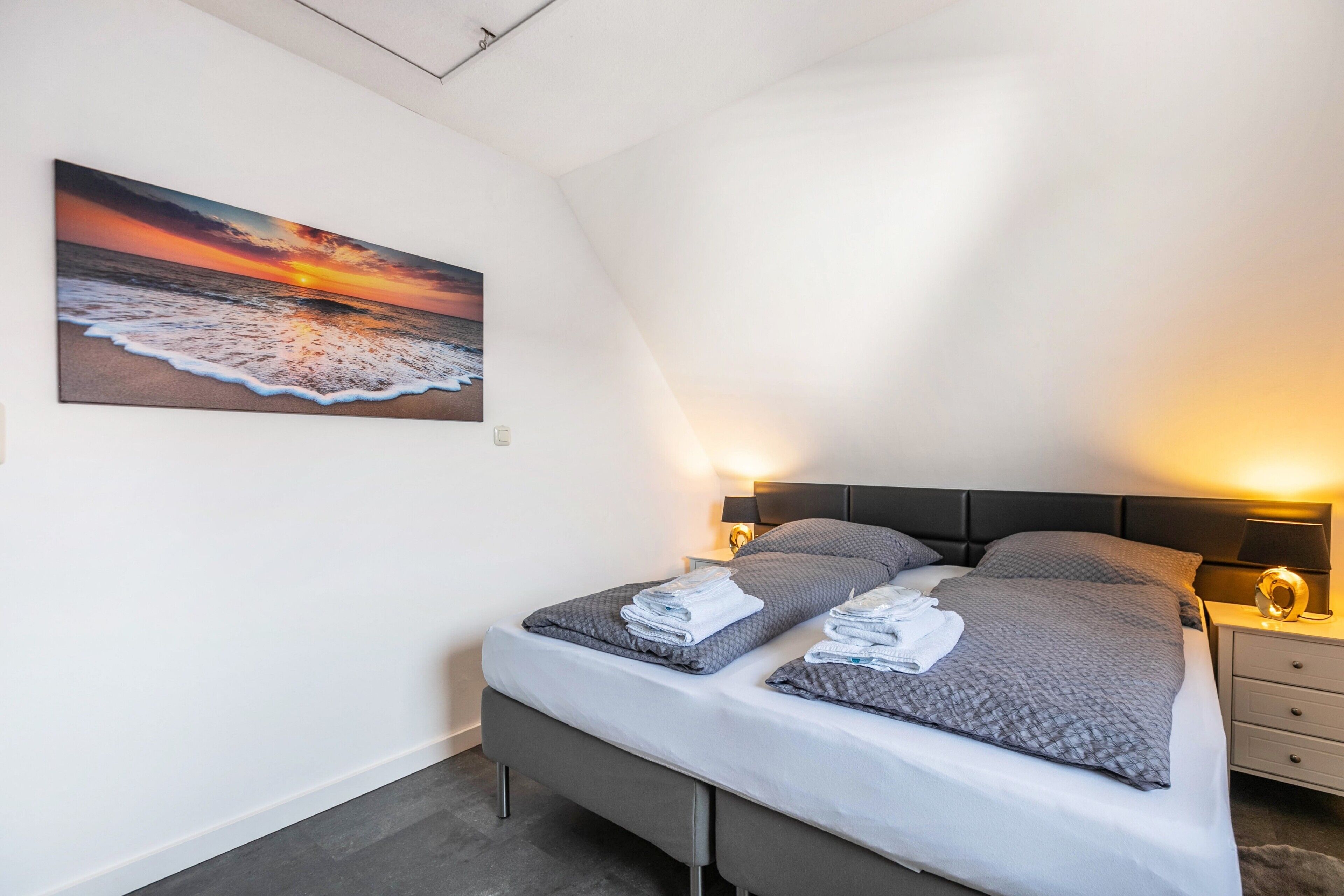 Ein Schlafzimmer mit einem Bett, Kissen, einem Nachttisch, einer Lampe und einem Sonnenuntergangsbild.