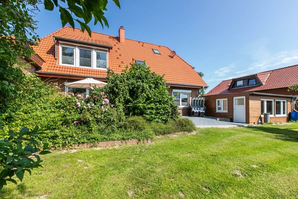A house with a red-tiled roof, a garden with pink flowers, and a clear blue sky.