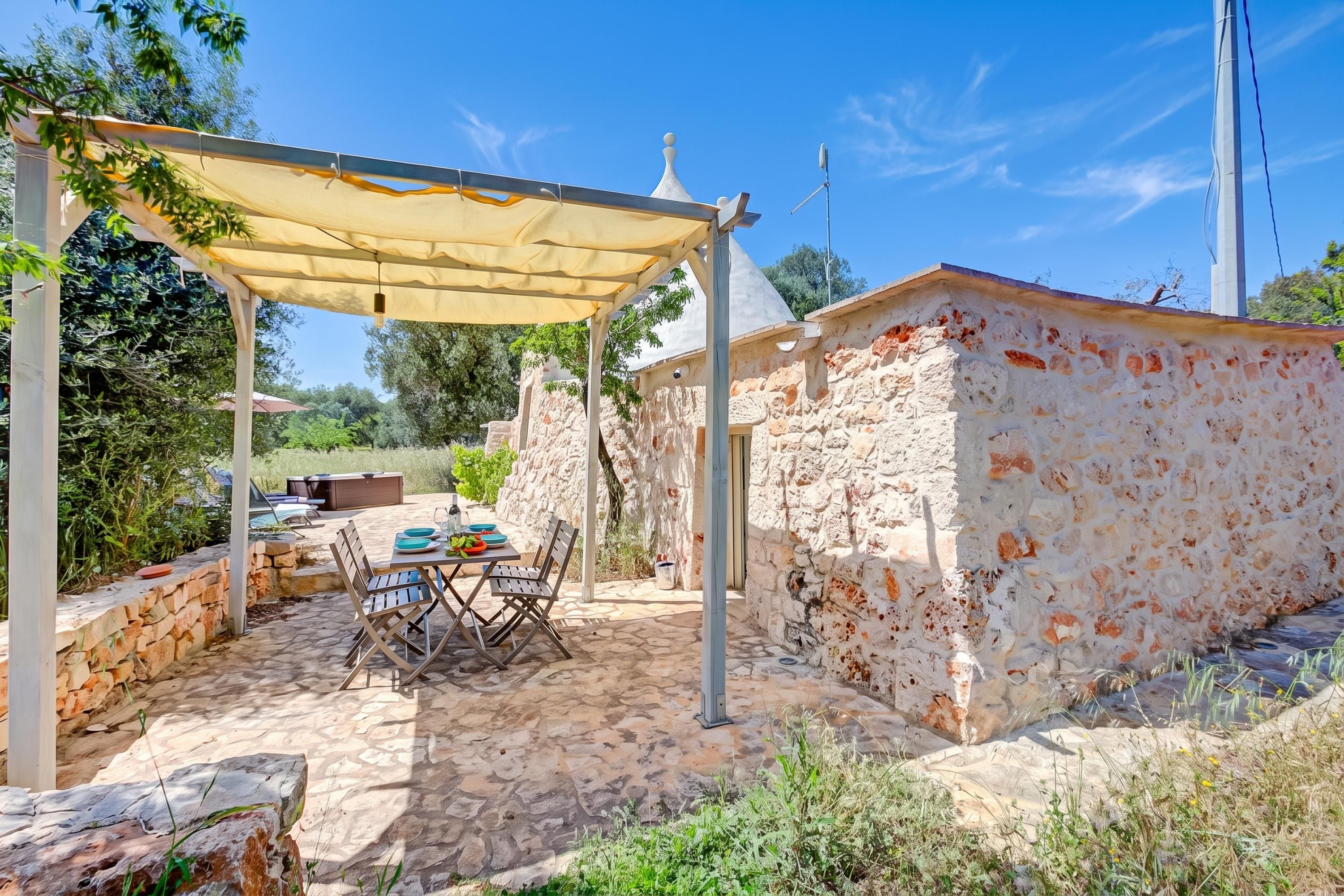 An outdoor patio area with a table and chairs, a pergola providing shade, and a stone wall.