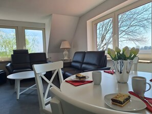 A modern room with a dining table, two chairs, a sofa, a lamp, and a window with a view of trees.