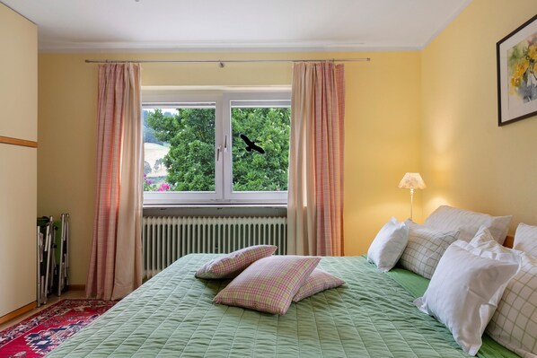 A bedroom with a green bedspread, checkered pillows, a radiator, a window with curtains, a lamp, and a framed picture on the wall. - Apartment 'Kneissler' with Mountain View, Private Terrace and Wi-Fi (Oberried)