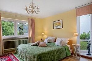 A bedroom with a green bedspread, two bedside lamps, a radiator, a window with curtains, and a painting on the wall.