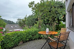 A patio with a round wooden table and chairs, surrounded by lush greenery and flowering plants.