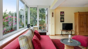 A living room with a sofa, a television, and a view of a garden through large windows. - Apartment 'Kneissler' with Mountain View, Private Terrace and Wi-Fi (Oberried)