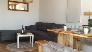 A modern living room with a wooden ceiling, a grey sofa, a white coffee table, and a wooden dining table with a furry bench. - Hestihof – Icelandic Horse Farm and Holiday Apartment (Waldburg)