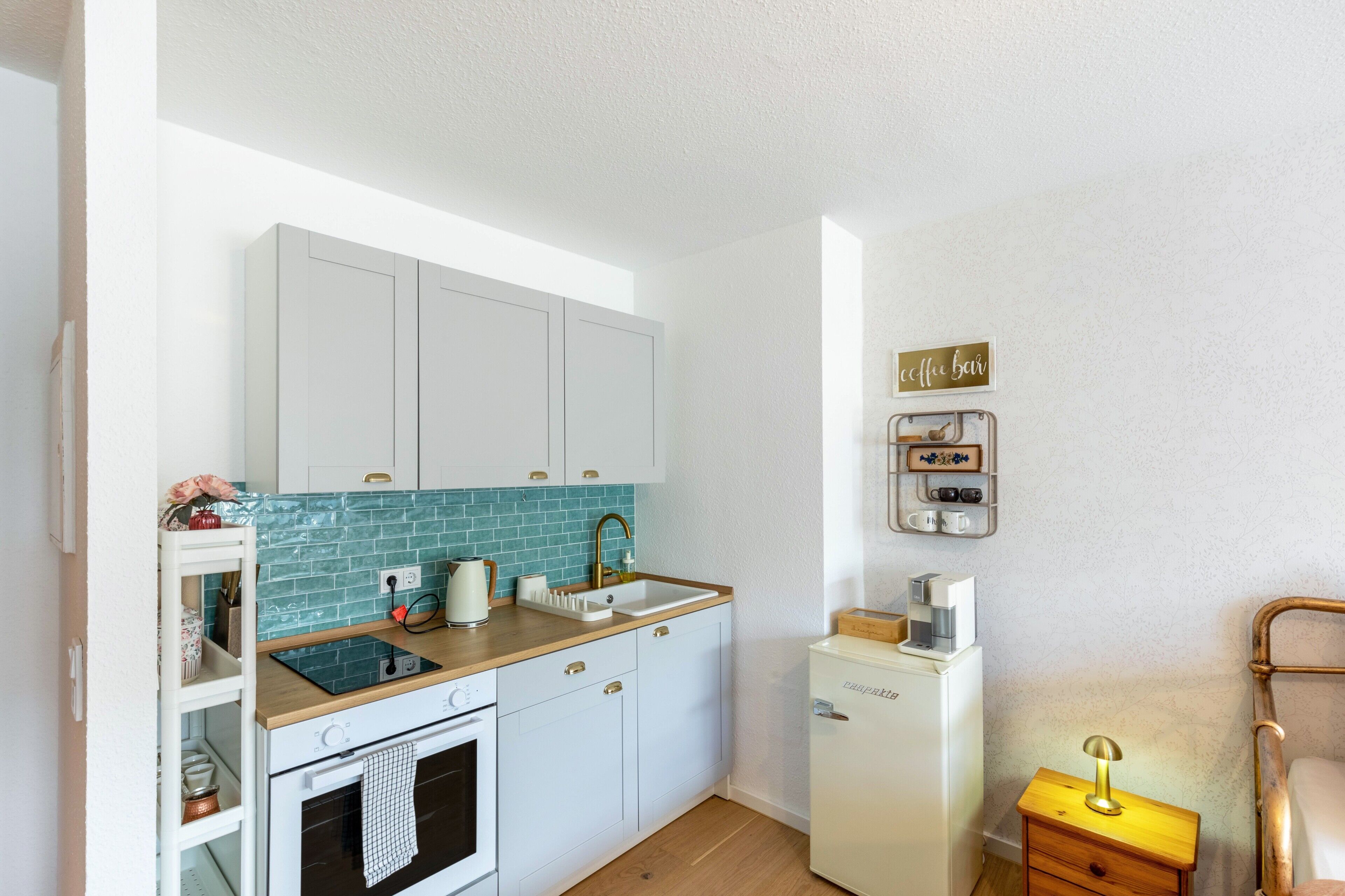 Une cuisine compacte comprenant un évier, une cuisinière et des placards, un petit réfrigérateur, ainsi qu’une table de chevet avec une lampe.