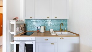Una cocina compacta con armarios blancos, salpicadero de azulejos azules, un fregadero, una estufa y una encimera con una tetera y una cafetera.