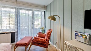 Una habitación con una silla de mimbre, un banco, una lámpara de pie y un televisor de pantalla plana montado en la pared.