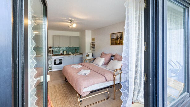 Un dormitorio con una cama, una mesita de noche, una ventana con cortinas, y una cocina al fondo, además de un ventilador de techo.