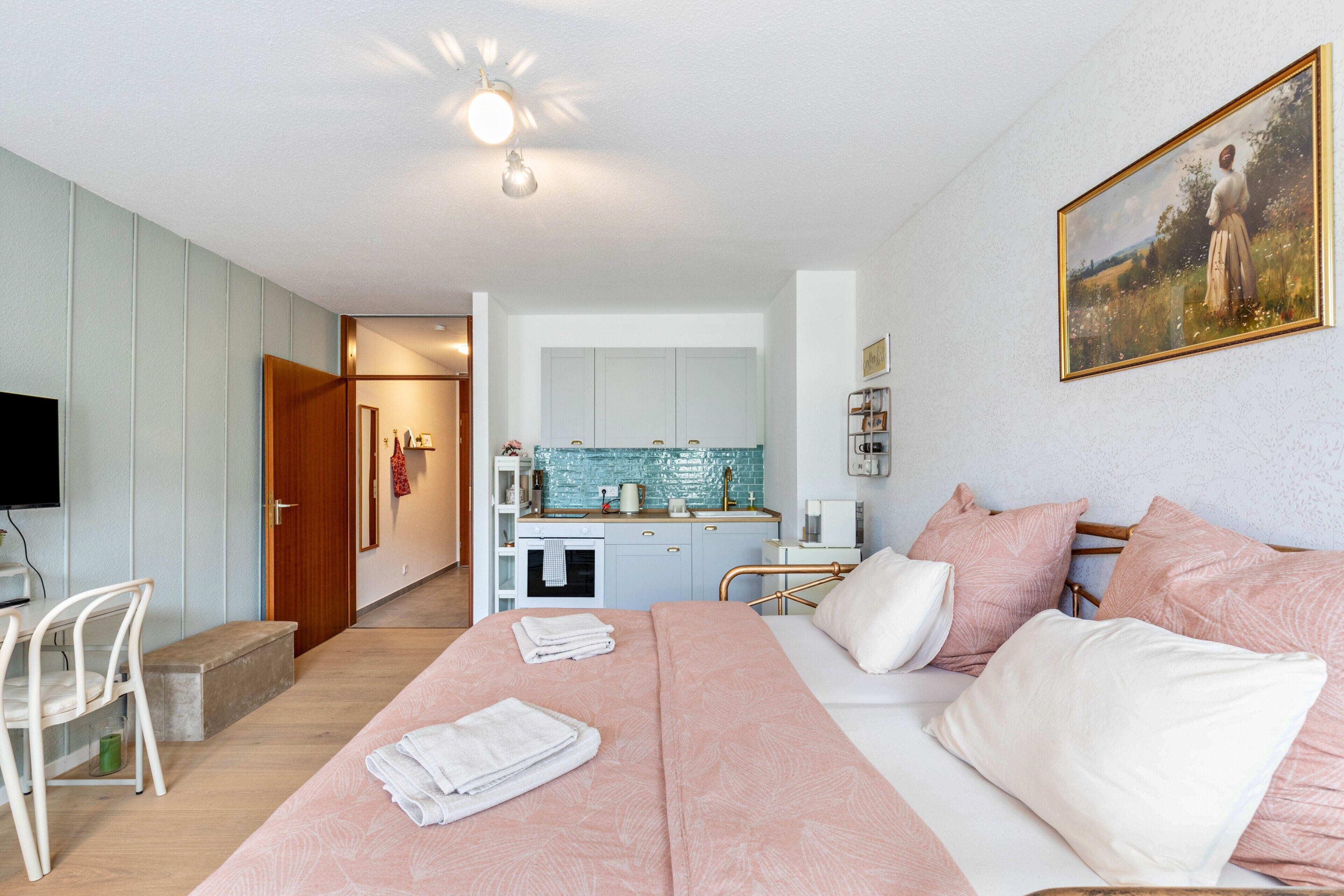 A bedroom with a bed, pillows, a bedside table, a TV, and a kitchen area with cabinets and a sink.