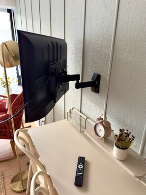 A wall-mounted TV with a remote control on a table, a clock, and a potted plant.