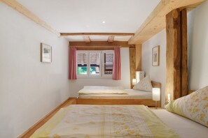 A bedroom with two beds, wooden beams, a window with shutters, and wall-mounted lamps.
