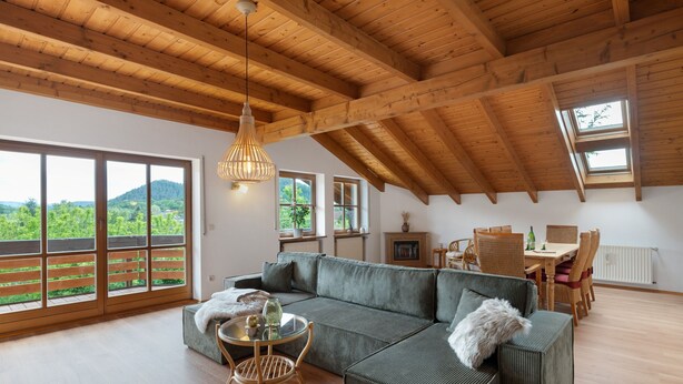 Un loft spacieux avec de hauts plafonds, de grandes fenêtres, un canapé d’angle, une table à manger et une vue sur la verdure.