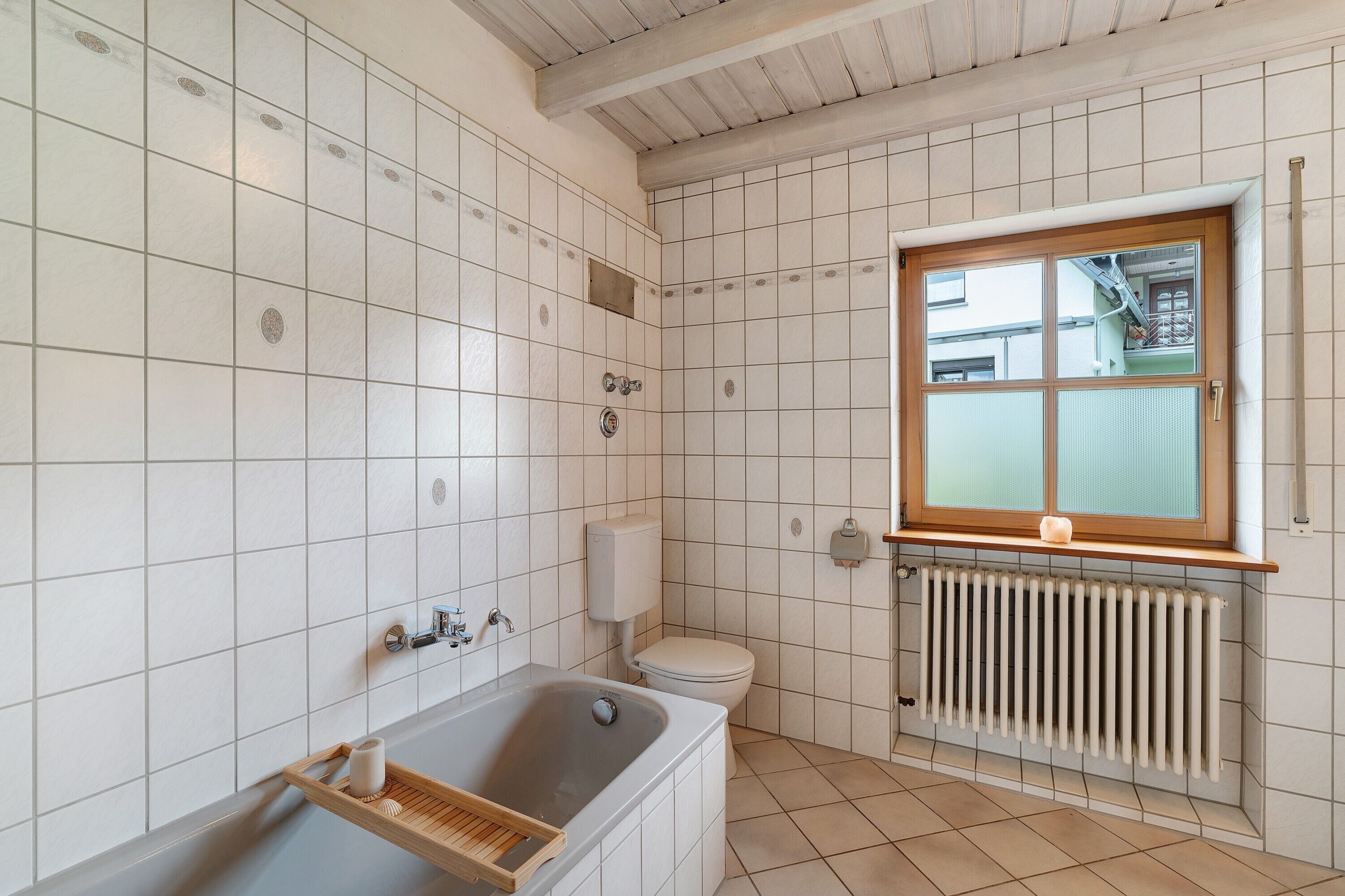 Un baño con paredes de azulejos blancos, una ventana de madera, una bañera, un inodoro y un radiador.