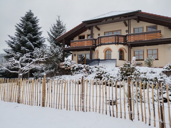 A two-story building with a balcony, surrounded by snow-covered trees and a wooden fence.
