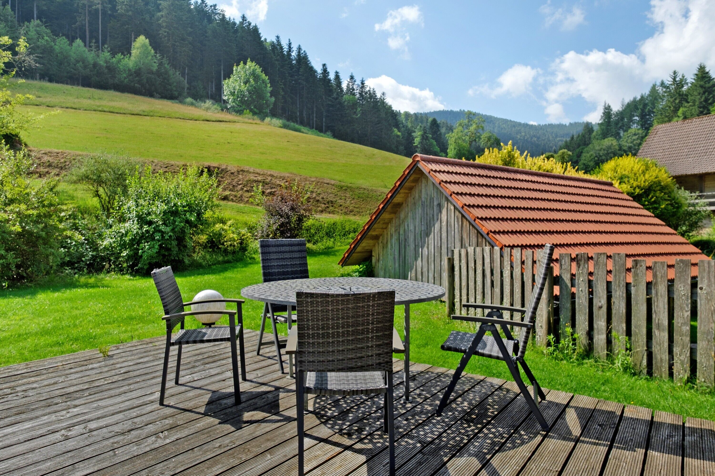 Une terrasse en bois avec des meubles d’extérieur, un petit abri de jardin et un paysage verdoyant, avec des arbres et des collines en arrière-plan.