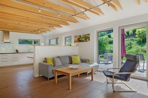 Ein modernes Wohnzimmer mit Sofa, Couchtisch aus Holz und Schaukelstuhl. Eine Küchenzeile mit weißen Schränken und Arbeitsplatte ist ebenfalls vorhanden. Große Fenster bieten Blick ins Grüne.