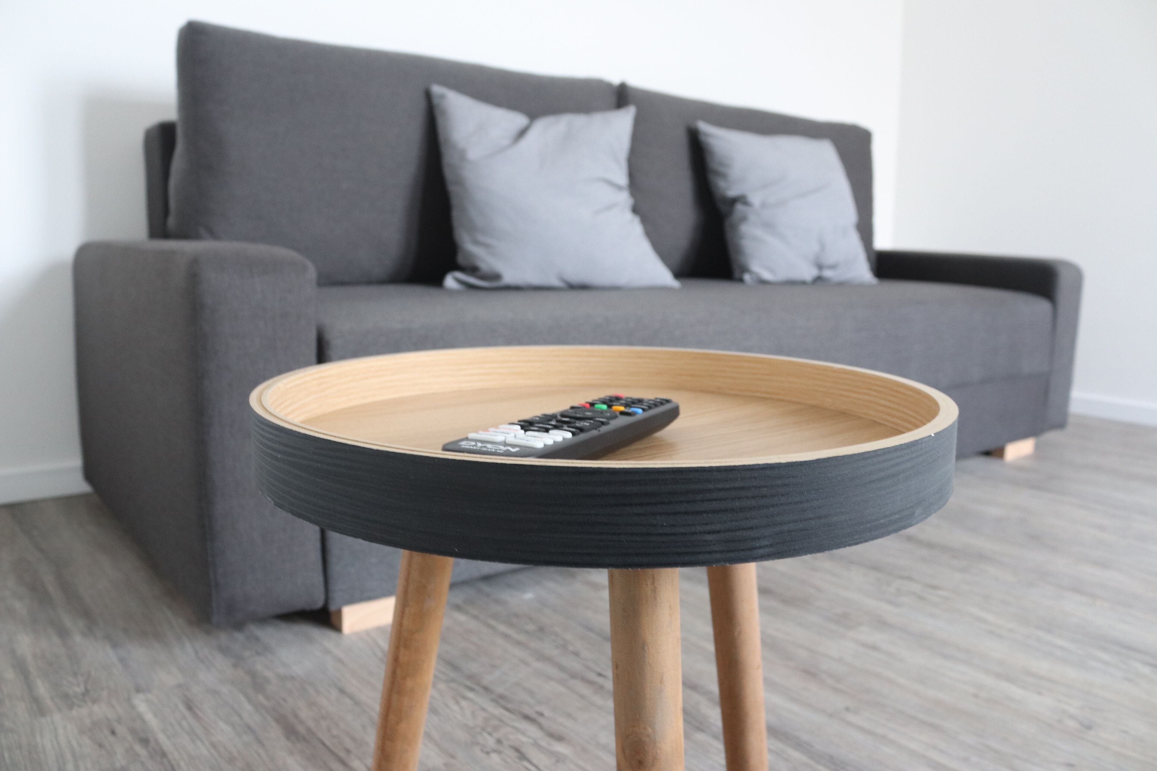 Una mesa de madera redonda con un control remoto encima, ubicada frente a un sofá gris con cojines.