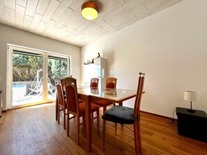 A dining area with a glass table, wooden chairs, a ceiling light, and a view through a glass door.