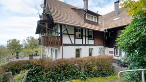 Ett traditionellt hus med korsvirkesfasad, ett sluttande tak, en balkong och en trädgårdsdel med tät buskvegetation.