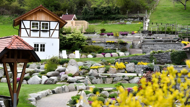 A two-story building with a wooden facade and a red roof, surrounded by a landscaped garden with rocks and flowers. - Apartment 'Mühlenstüble' with Mountain View, Shared Terrace and Wi-Fi (Gutach (Schwarzwaldbahn))