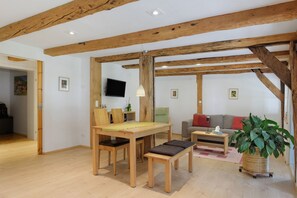 Een kamer met houten balken, een eettafel met stoelen, een bank, een televisie en een potplant.