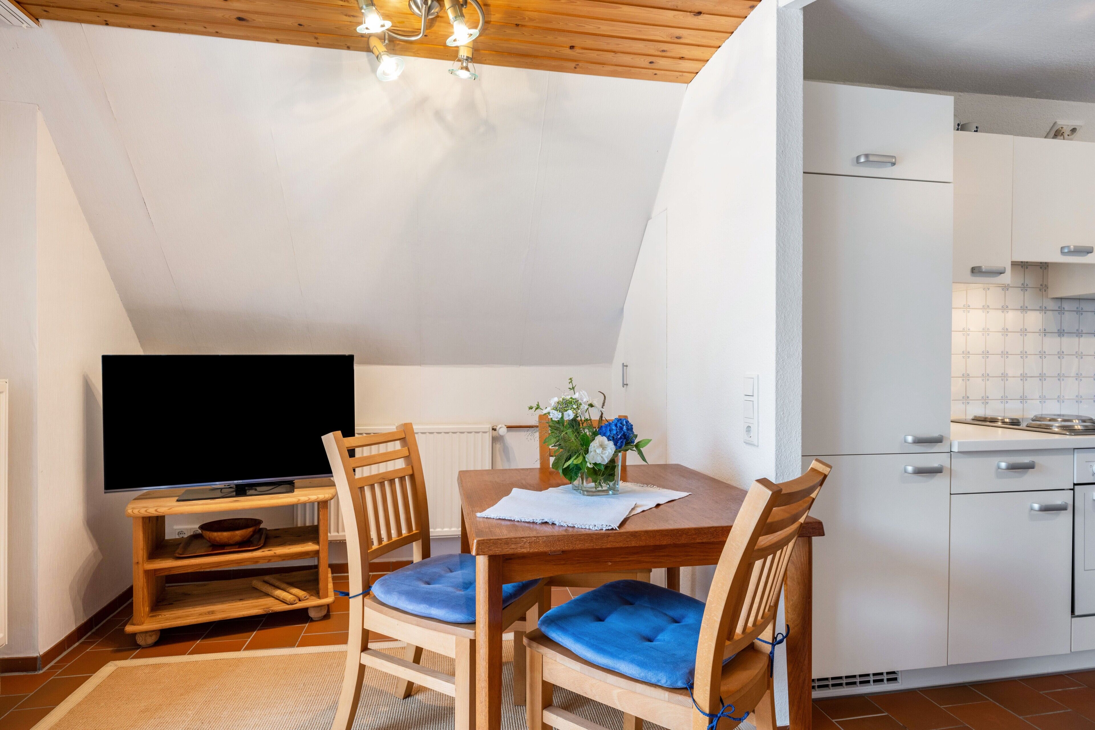 En matplats med en tv, trästolar med blå kuddar, ett litet bord med en vas med blommor och ett kök med vita skåp.