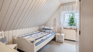 A bedroom with a sloped ceiling, a bed with a blue and white bedspread, a bedside table with a lamp, and a window with a view of greenery. - Apartment 'Ella Walter, Kiek Ut' with Private Terrace, Shared Garden and Wi-Fi (Nordstrand)