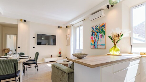 A modern kitchen with a large island, a dining area with a green upholstered chair, and a colorful painting on the wall.