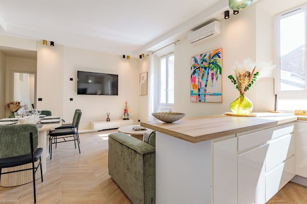 A modern kitchen with a large island, a dining area with a green upholstered chair, and a colorful painting on the wall.