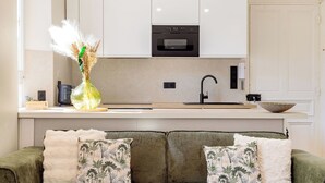 A modern kitchen with a built-in oven, a sink, and a countertop. There is a green sofa with floral cushions in the foreground.