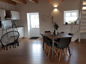 Un comedor con una mesa y sillas de madera, un sillón colgante, una cocina con armarios blancos y una escalera.