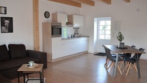 Una cucina moderna con mobili bianchi, forno integrato e finestra. Nell'area soggiorno, un tavolo da pranzo con sedie e un divano di colore scuro.