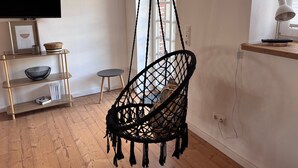 A room with a hanging chair, a TV, a shelf with decorative items, and a window with a view. - Apartment 'Wanderlust' with Private Terrace, Private Garden and Wi-Fi (Heide)