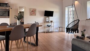 A modern living room with a wooden dining table, grey chairs, a hanging chair, a TV, and a kitchen area with an oven. - Apartment 'Wanderlust' with Private Terrace, Private Garden and Wi-Fi (Heide)