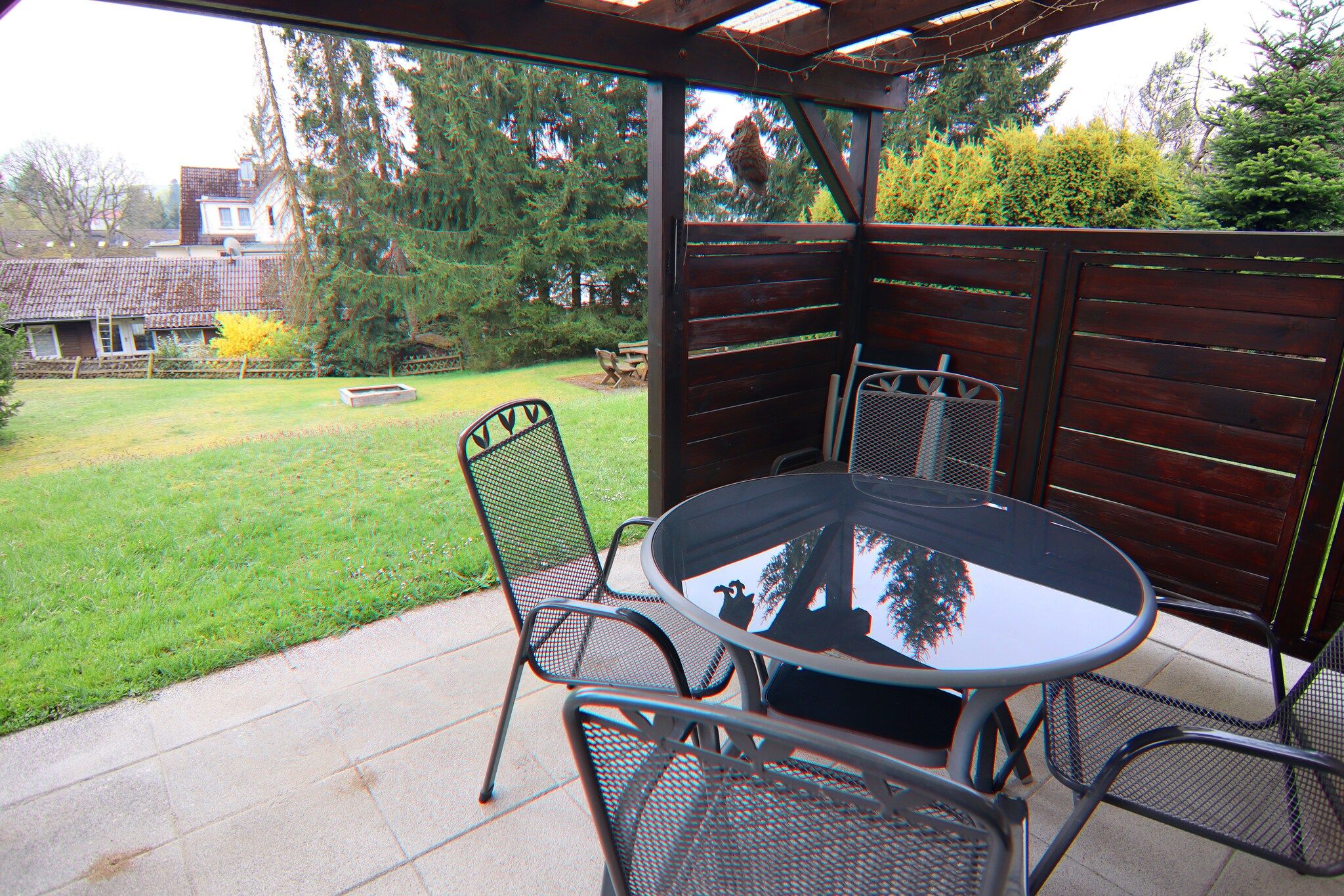 Un patio cubierto con una mesa redonda y cuatro sillas, una valla de madera, un césped con árboles y una casa al fondo.