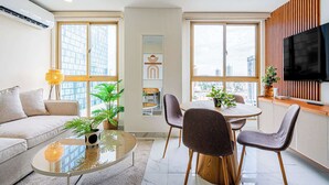 Ein modernes Wohnzimmer mit Sofa, Couchtisch und Essbereich mit Stühlen. Ein Fernseher ist an der Wand befestigt, eine Topfpflanze steht dort und es gibt ein Fenster mit Blick auf die Stadt.
