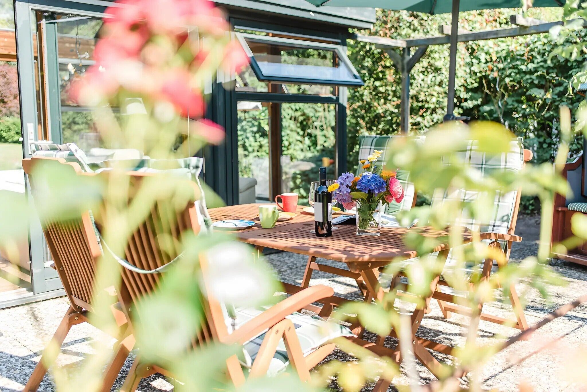 Un espace de restauration en plein air avec des meubles en bois, une table ornée d’une bouteille et de fleurs, et une structure vitrée abritant des plantes.