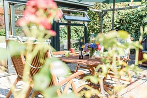 An outdoor dining area with wooden furniture, a table with a bottle and flowers, and a glass enclosure with plants.
