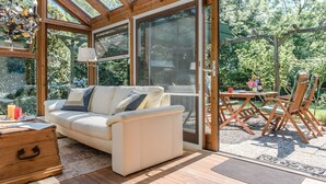 A sunlit room with a sofa, wooden drawers, and a view through large glass doors to an outdoor area with a table and chairs. - Holiday Home 'Am Meer' with Sea View, Private Terrace and Wi-Fi (Langballig)
